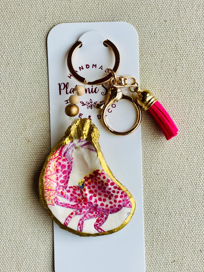 May include: Handmade oyster shell keychain with a gold-painted edge and a pink crab design. The keychain has a gold-colored ring, a pink tassel, and wooden beads. The background is a white card with text.