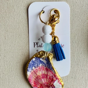 May include: A handmade keychain featuring a painted oyster shell with a gold-painted edge. The shell is decorated with red, white, and blue patterns. The keychain includes white and blue beads, a blue tassel, and a gold-colored clasp.
