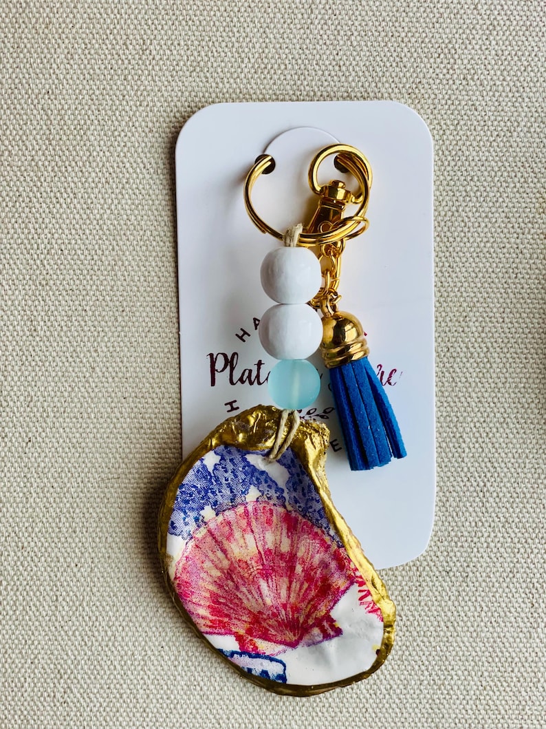 May include: A handmade keychain featuring a painted oyster shell with a gold-painted edge. The shell is decorated with red, white, and blue patterns. The keychain includes white and blue beads, a blue tassel, and a gold-colored clasp.