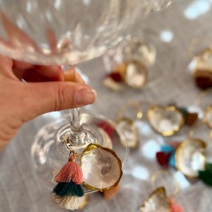May include: A set of wine glass charms featuring small, polished oyster shells with gold-painted edges, each paired with a multi-colored tiered tassel in shades of pink, teal, and cream, attached to a thin gold-toned metal hoop ring.