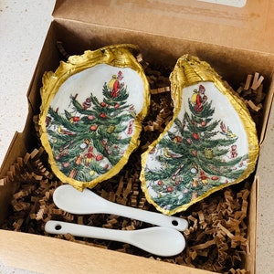 May include: Two white oyster shells with gold trim, each painted with a Christmas tree and a Santa Claus figure. The shells are in a brown box with shredded paper. Two white spoons are also included.