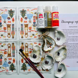 May include: A DIY decoupage kit for oyster shells with instructions, white acrylic paint, gold paint, Mod Podge, and two paintbrushes. The kit includes six oyster shells, each with a different design, and a sheet of paper napkins with a Nutcracker theme.