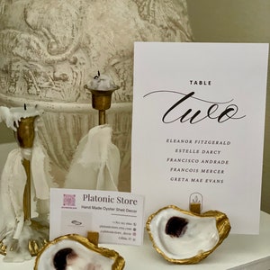 May include: A white oyster shell with gold trim, used as a table number holder. The table number is "Two" and the names of the guests are listed below. The oyster shell is sitting on a table with a business card for "Platonic Store" in the background.