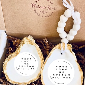 May include: Two oyster shells with gold-painted edges and white centers, each featuring a circular label with the text 'YOUR LOGO OR CUSTOM PICTURE'. One shell includes a white beaded loop with a ribbon. The items are inside a cardboard box.