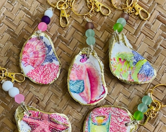 May include: Six oyster shell keychains with gold accents and colorful painted designs. Each keychain features a different sea creature, including a starfish, a seahorse, and coral. The keychains are strung with colorful beads and gold chains.
