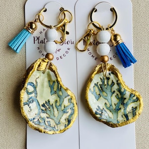May include: Two oyster shell keychains with gold-painted edges and blue coral designs. Each keychain has a gold clasp, white beads, a gold bead, and a blue tassel. The keychains are displayed on white cards with text.