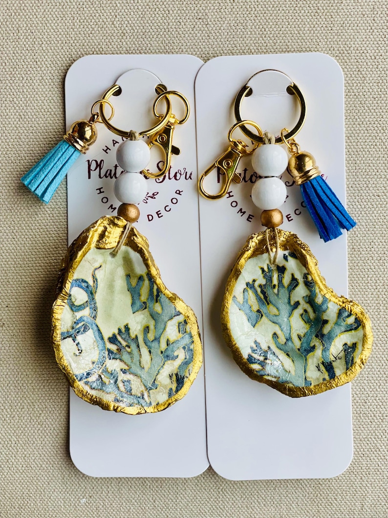May include: Two oyster shell keychains with gold-painted edges and blue coral designs. Each keychain has a gold clasp, white beads, a gold bead, and a blue tassel. The keychains are displayed on white cards with text.