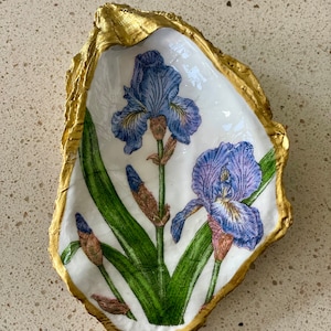 May include: A white oyster shell with a gold rim, painted with a blue and purple iris flower design.