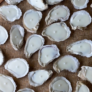 May include: A collection of white oyster shell candles arranged on a woven tray. The candles are all lit and the wax is melted, creating a warm and inviting atmosphere.