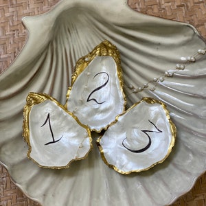 May include: Three white oyster shells with gold trim, each with a black number written inside. The shells are arranged in a larger white seashell with a gold trim.
