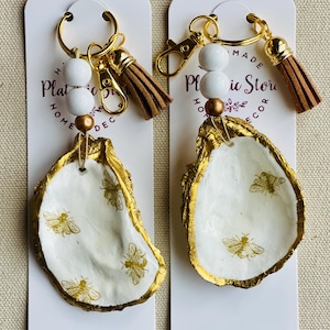 May include: Two keychains featuring white oyster shells with gold edges and bee designs. Each keychain has a gold ring, white beads, a gold bead, and a brown tassel. The keychains are displayed on white cards with text.