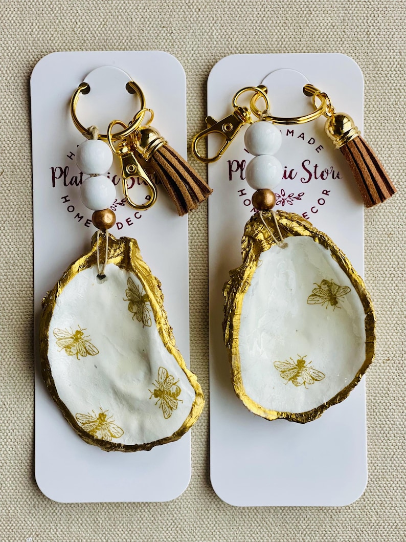 May include: Two keychains featuring white oyster shells with gold edges and bee designs. Each keychain has a gold ring, white beads, a gold bead, and a brown tassel. The keychains are displayed on white cards with text.
