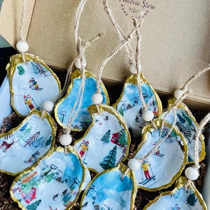May include: A set of 10 oyster shell ornaments with a winter wonderland scene painted on each. The shells are painted with gold trim and have a white wooden bead at the top of each string.