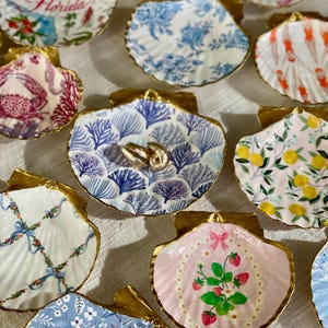 May include: A collection of decorative seashell dishes with gold-painted edges. Each shell features a unique hand-painted design, including floral patterns, coral, and illustrations of Florida. Ideal for holding jewelry or small items.
