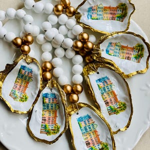 May include: A collection of decorative oyster shells with gold-painted edges, each featuring a watercolor painting of a colorful building. White and gold beaded necklaces are also present, creating a coastal theme.