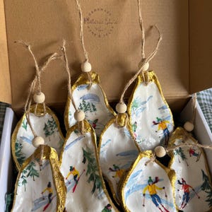 May include: A collection of handmade oyster shell ornaments. Each shell is hand-painted with a winter ski scene and trimmed with gold paint. The ornaments are strung with twine and a wooden bead. The box reads "HANDMADE Platonic Store HOME & DECOR".