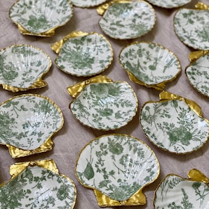 May include: A collection of decorative seashell-shaped dishes. Each dish features a white background with a green floral pattern and a gold-colored base. The dishes are arranged on a neutral-colored surface.