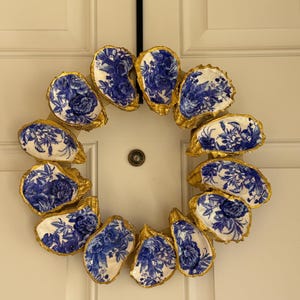 May include: A decorative wreath made from oyster shells. Each shell is painted with a blue floral design and trimmed with gold. The wreath is hanging on a white door.