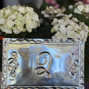 May include: A rectangular, silver-colored metal plaque with a decorative border and the embossed number "2". The plaque features floral designs on the corners. White hydrangea flowers are in the background.