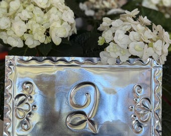 May include: A rectangular, silver-colored metal plaque with a decorative border and the embossed number "2". The plaque features floral designs on the corners. White hydrangea flowers are in the background.