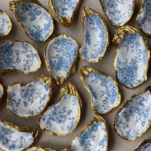 May include: A collection of decorative oyster shells. Each shell features a white interior with a blue floral pattern and a gold-painted rim. The shells are arranged on a neutral-colored surface.