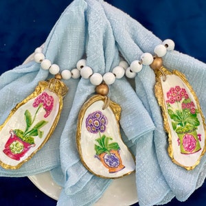 May include: Three oyster shell napkin rings with gold trim and painted floral designs. Each ring features a different flower in a pot, with pink, purple, and green colors. The rings are strung on a white beaded chain.