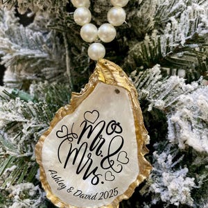 May include: A decorative oyster shell ornament with gold edging, strung with a pearl necklace. The shell features the words "Mr. & Mrs. Ashley & David 2025" in elegant script, perfect for wedding decor.