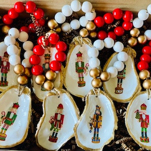 May include: Six oyster shell ornaments with gold trim, each featuring a different nutcracker design. The ornaments are surrounded by red and white beaded garlands.