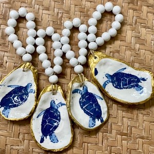 May include: Four oyster shell ornaments with a white base, gold trim, and a blue sea turtle design. Each shell is strung on a white beaded strand, perfect for coastal decor.