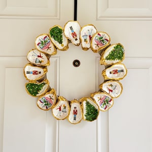 Puede incluir: Una corona navideña hecha con conchas de ostra. Cada concha tiene un borde dorado y presenta un diseño festivo, incluyendo árboles de Navidad y cascanueces. La corona está colgada en una puerta blanca.