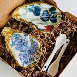 May include: Two small, white ceramic oyster shells with gold trim. The shells are decorated with floral patterns. One shell has a blue iris pattern, the other has a purple hyacinth pattern. Two small white ceramic spoons are included.