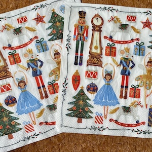 May include: Two white decorative napkins with a Christmas theme. Each napkin features illustrations of a nutcracker, clock, Christmas tree, ballerina, and wrapped gifts in red, blue, green, and gold.