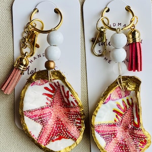 May include: Two handmade keychains featuring painted oyster shells with a gold trim and a starfish design in shades of pink and red. Each keychain has a gold-colored clasp, white beads, and a pink tassel. The text "HANDMADE" is visible.