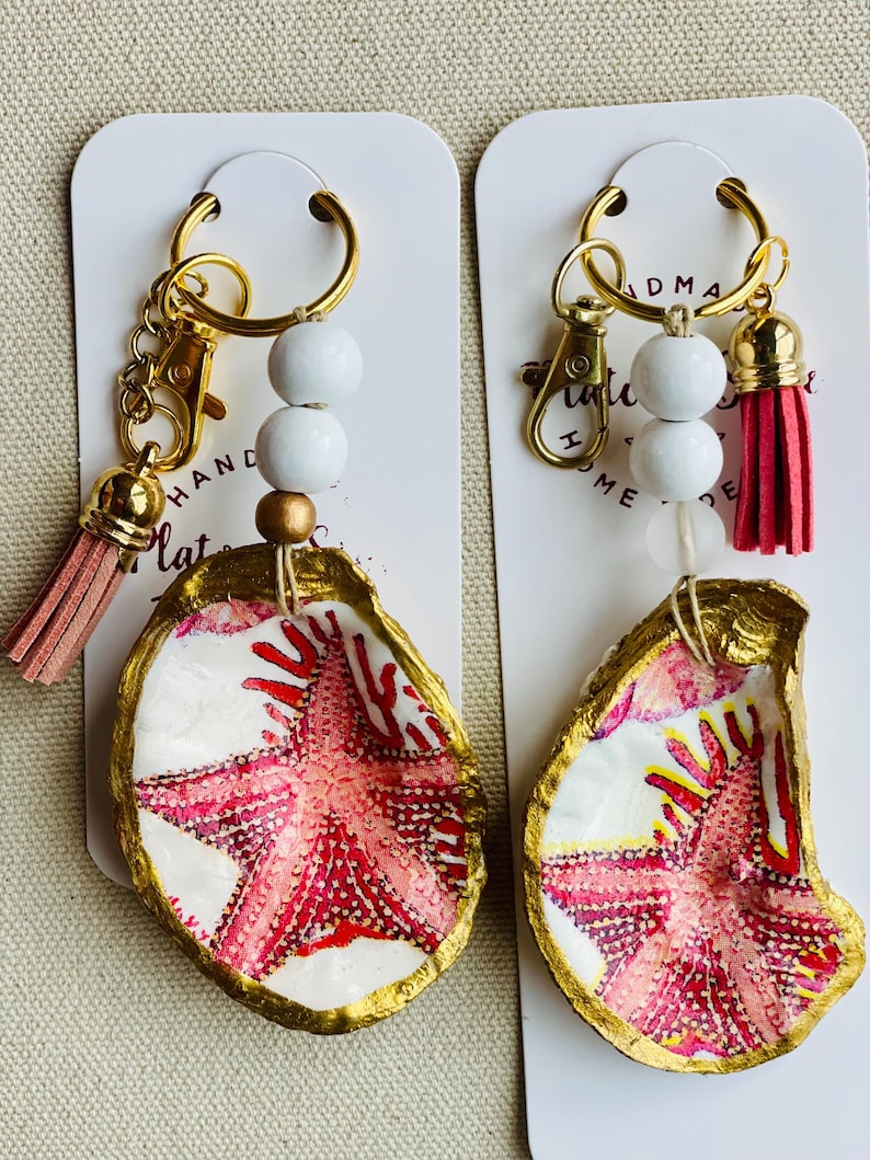 May include: Two handmade keychains featuring painted oyster shells with a gold trim and a starfish design in shades of pink and red. Each keychain has a gold-colored clasp, white beads, and a pink tassel. The text "HANDMADE" is visible.