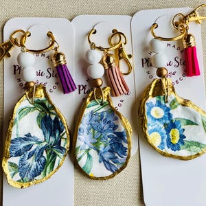 May include: Three oyster shell keychains with gold-colored trim and hardware. Each shell features a different floral design in blue and white. The keychains have white beads and a tassel in purple, pink, and tan.