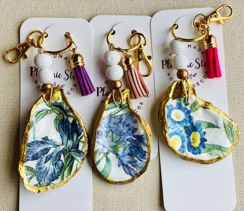 May include: Three oyster shell keychains with gold-colored trim and hardware. Each shell features a different floral design in blue and white. The keychains have white beads and a tassel in purple, pink, and tan.
