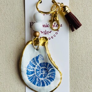 May include: Key chain featuring a white and gold painted oyster shell with a blue spiral design. The key chain has a gold clasp, white wooden beads, and a brown tassel. The tag reads "nic store decor".