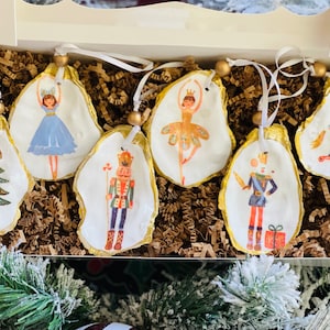 May include: Six Christmas ornaments shaped like oyster shells with gold trim. The ornaments feature painted images of a Christmas tree, a ballerina, a nutcracker, and a rocking horse.