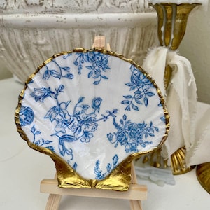 May include: A seashell-shaped decorative plate with a gold-painted rim, resting on a small wooden easel. The plate features a white background with blue floral patterns. The gold rim adds a touch of elegance to the design.