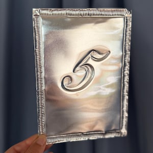 May include: A rectangular silver-colored metal sign featuring a raised, embossed number 5 in the center. The sign has a decorative, crimped border along all four edges, creating a textured frame around the smooth, reflective surface of the metal.