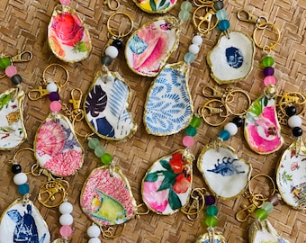 May include: A collection of 18 oyster shell keychains with various colorful designs and gold accents. Each keychain features a different pattern, including floral, geometric, and abstract designs. The keychains are strung with beads and attached to gold key rings.