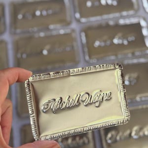 May include: A rectangular silver-toned metal plaque featuring a decorative crimped border and the embossed script name "Michell Wayne" centered on the surface. The plaque has a polished, reflective finish and is part of a collection of similar items.