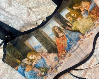 May include: A black corset top featuring a print of the Last Supper painting. The top has black straps and trim, with a lace-up detail on the side. The artwork is in muted tones of blue, red, and yellow.