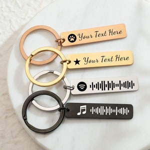 May include: Four personalised keyrings with metal rings. One rose gold keyring has a paw print and the text "Your Text Here". A gold keyring has a star and the text "Your Text Here". A silver keyring has a Spotify code. A black keyring has a musical note and a soundwave.