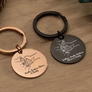 Op de afbeelding: Twee sleutelhangers met gegraveerde vuiststoten en namen. De sleutelhanger links is roségoud met de namen Amelia, Barbara, Olivier en Daddy. De sleutelhanger rechts is zwart met de namen William, Amelia, Barbara, Olivier en Daddy.