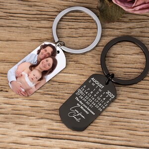 May include: Two keychains with metal rings. One keychain has a photo of a family. The other is black with the text "AUGUST 2023" and a calendar, with the text "The day you became my Mommy Love from Charlie."
