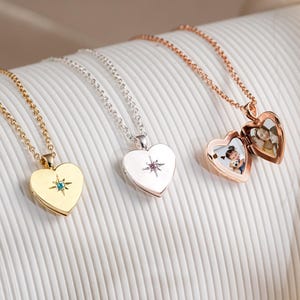 May include: Three heart-shaped lockets on delicate chains. One is gold with a blue gemstone, another is silver with a purple gemstone, and the third is rose gold, open to reveal two small photos. The lockets are displayed on a white, textured surface.