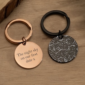 May include: Two keychains with round metal tags. One tag is rose gold with the text "The night sky on our first date x". The other tag is black with a white constellation design.