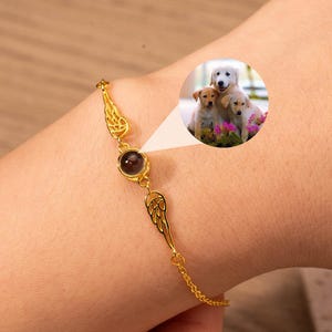 Puede incluir: Una pulsera dorada con una gema central de color rojo oscuro y delicados detalles en forma de alas. La pulsera se muestra en una muñeca, con una pequeña imagen insertada de tres cachorros.