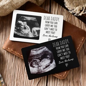 May include: Two rectangular wallet cards featuring a black and white ultrasound image and the text: Dear Daddy. Now you can carry me too. Can't wait to meet you! Love, the bump. One card is white with black text, and the other is black with white text.